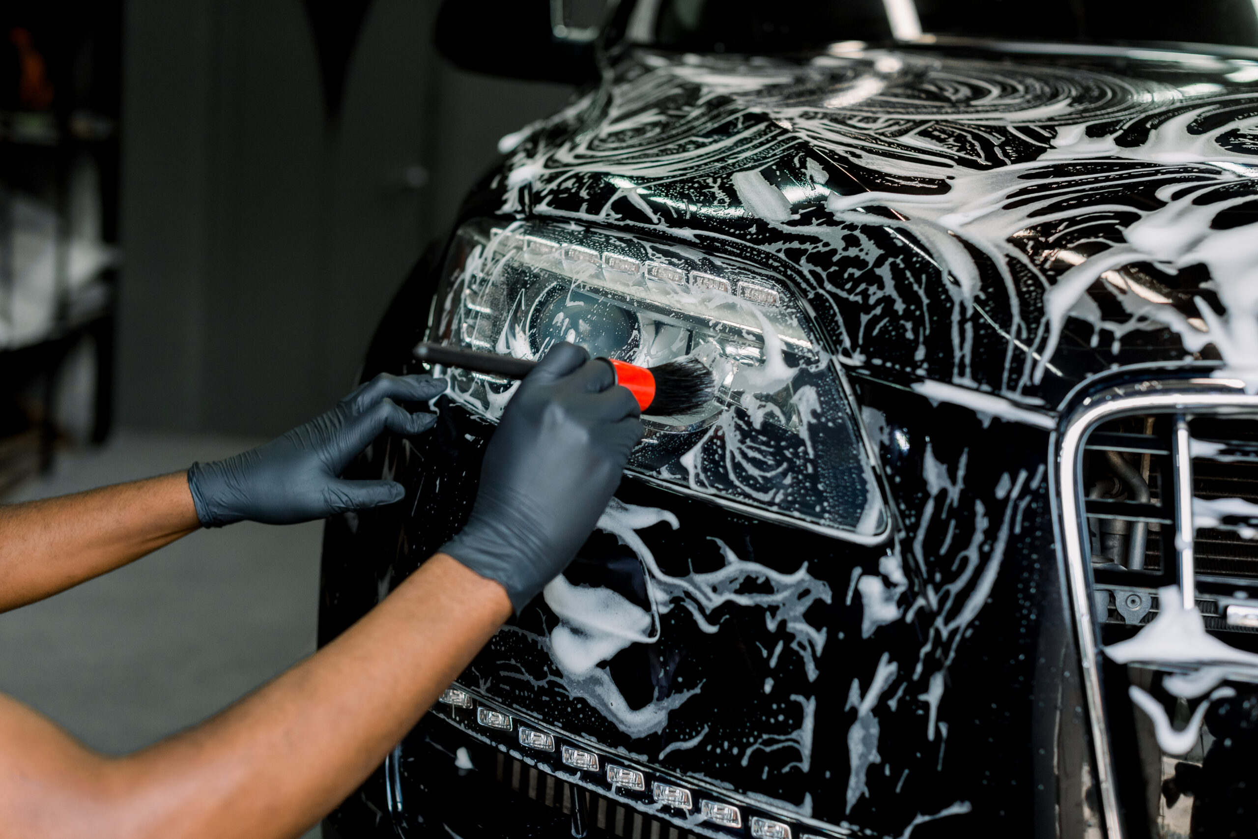 Close up photo of hands of auto service male worker in black protective gloves cleaning car hood and headlights with foam and soft brush. Carwash and detailing